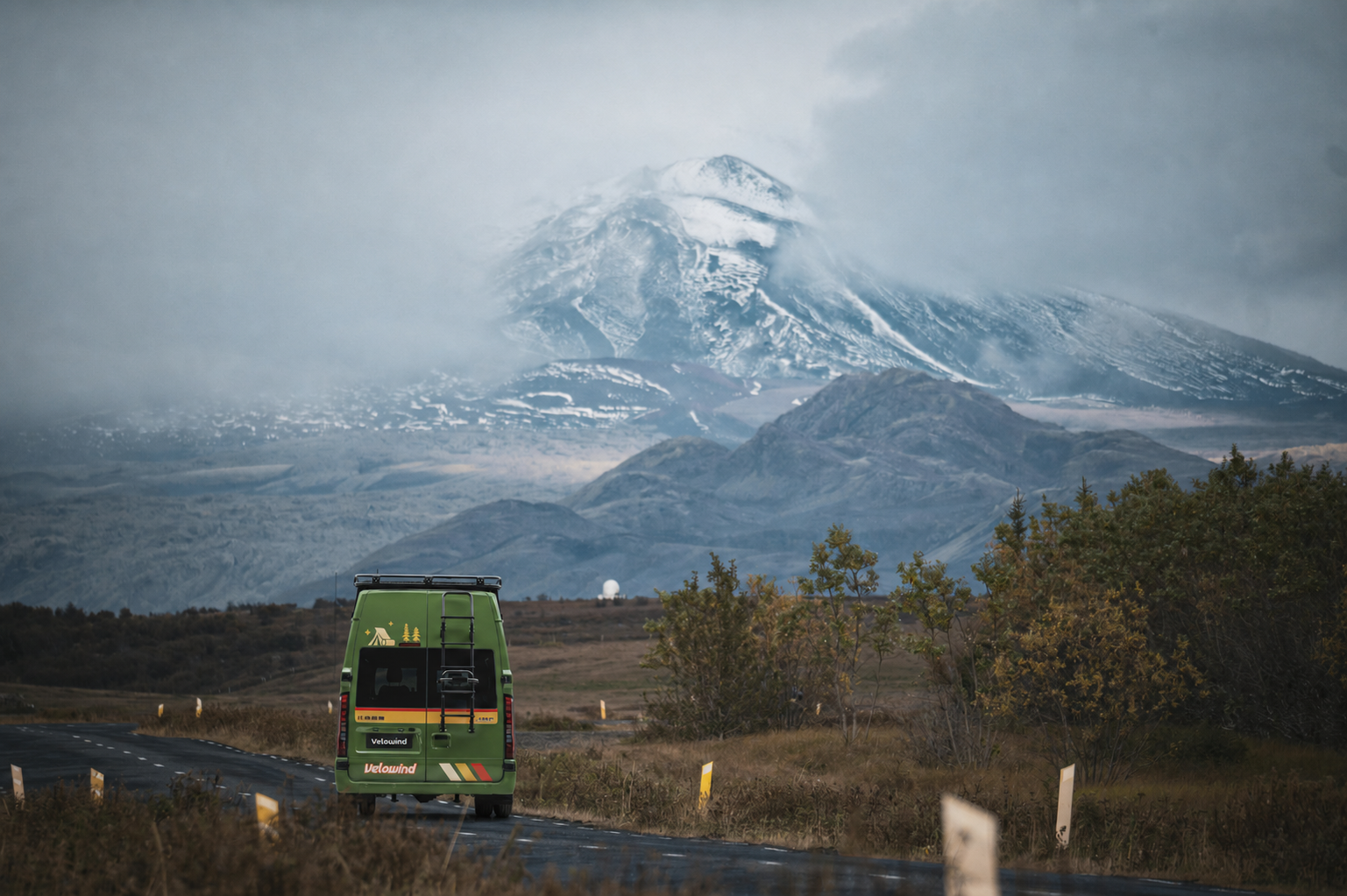 A green Velowind support vehicle heading toward a mountain road.